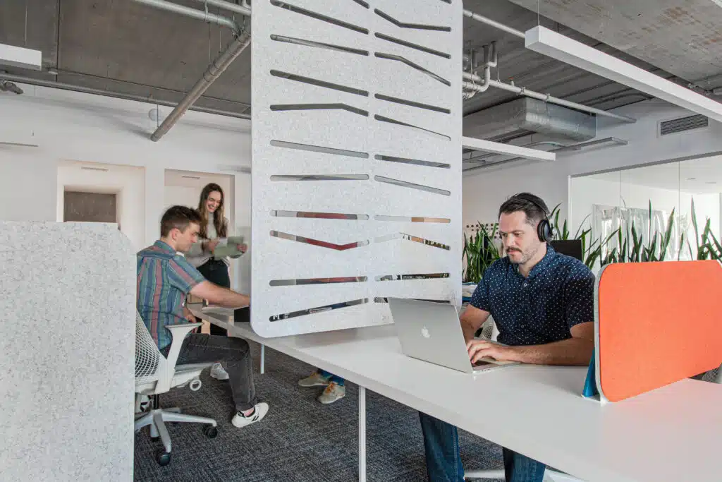 Modern open office with Loftwall acoustic divider and desk screens creating privacy zones; employees work and collaborate in a bright, flexible workspace.